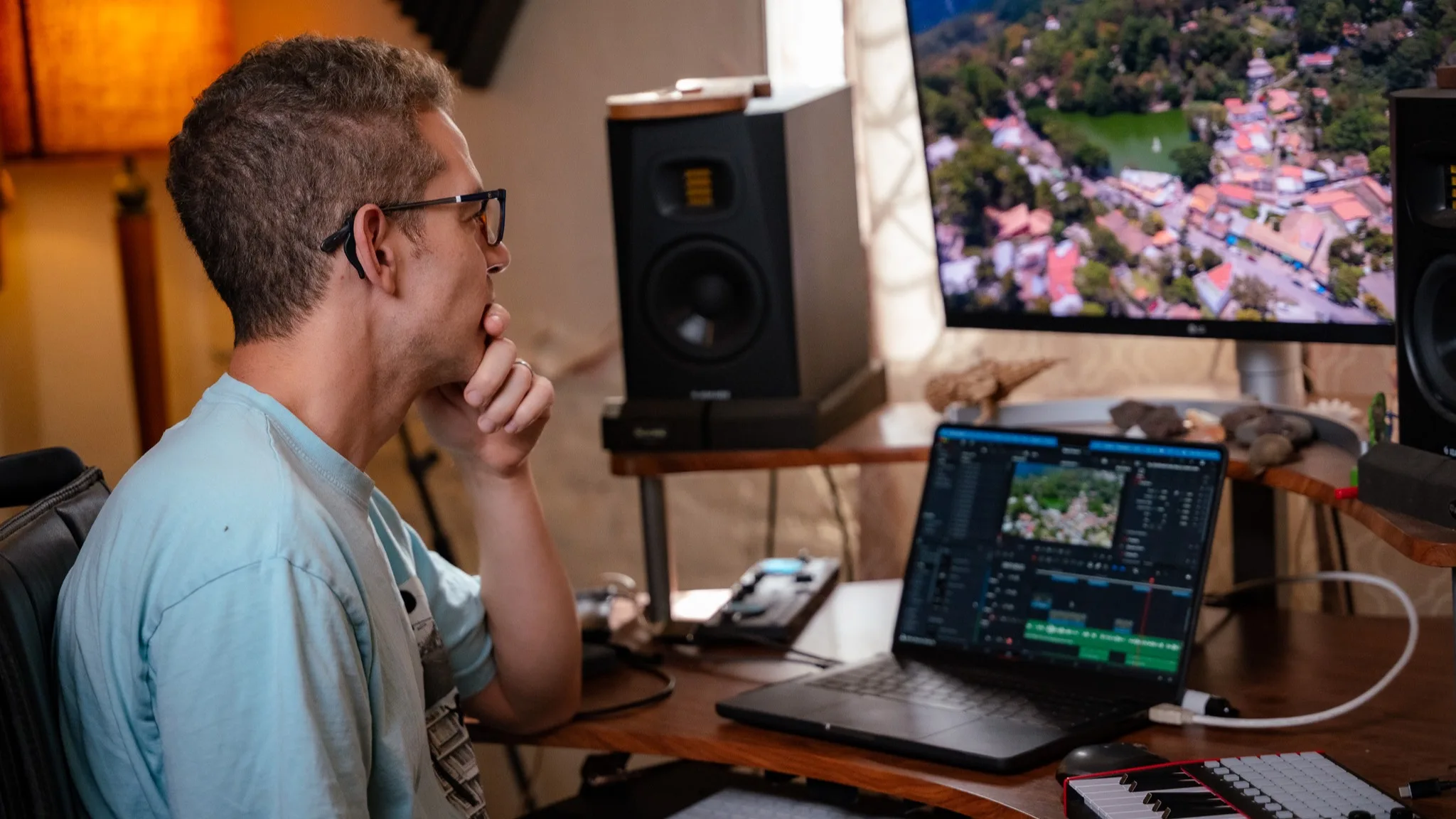 Juan Pablo Lagos at the studio desk, hand at his chin, watching an aerial shot play back on the monitor with the edit timeline open on the laptop beside him.